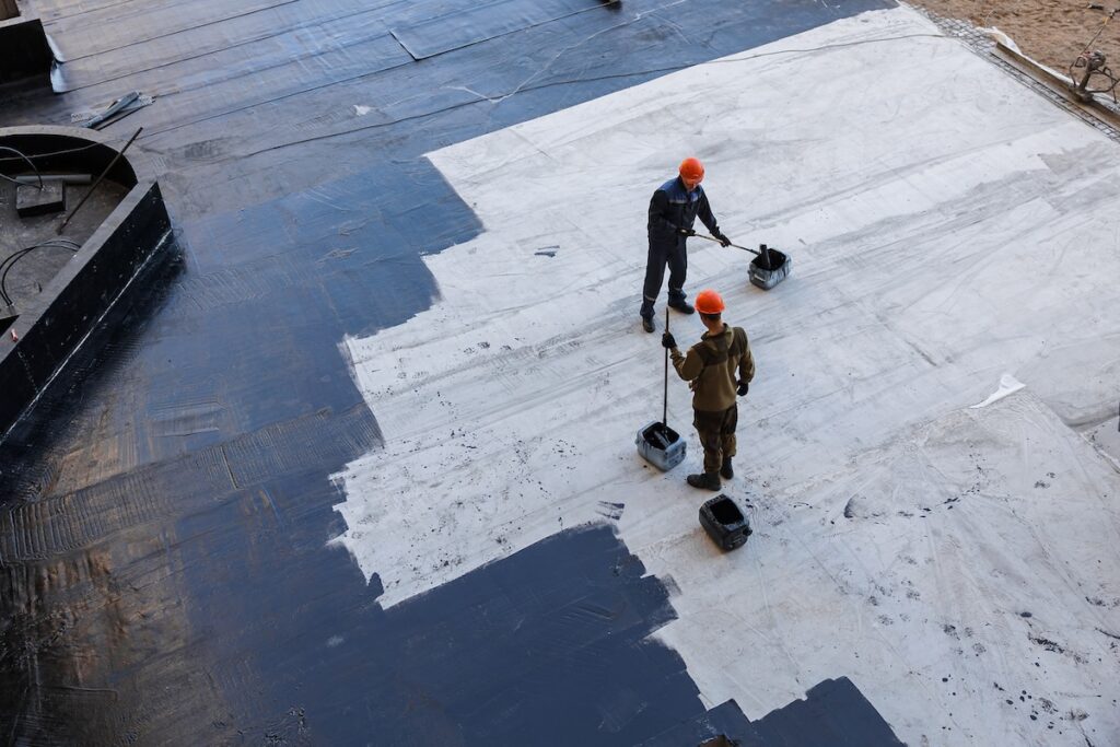 Two construction workers in orange helmets apply black waterproofing material to a large concrete surface using long-handled rollers.