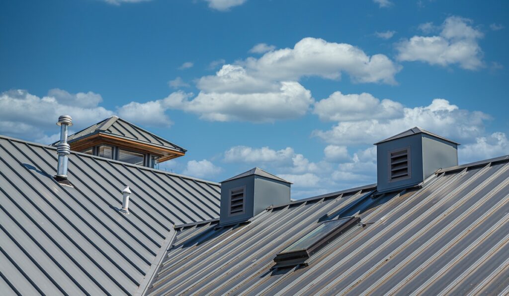 A close-up view of a black metal roof with two cupolas and a chimney vent under a blue sky filled with fluffy white clouds. The metal panels have diagonal lines, adding texture to the scene.