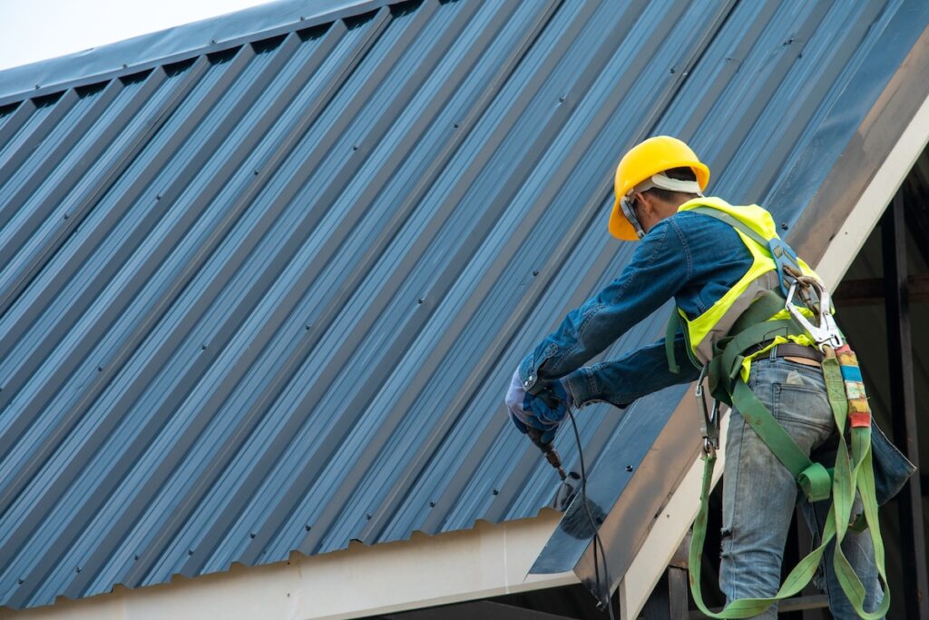 A worker in a yellow hard hat and safety harness is installing black metal roof.