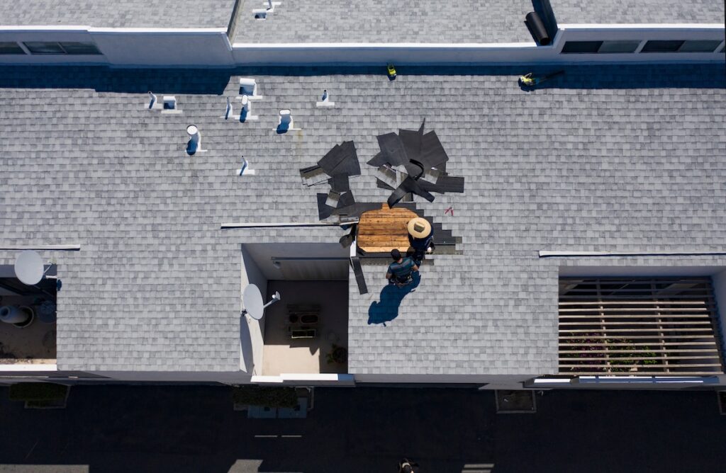 Aerial view of two people repairing shingles on a gray rooftop. Tools and replacement shingles are scattered around them, and their shadows are visible on the roof.