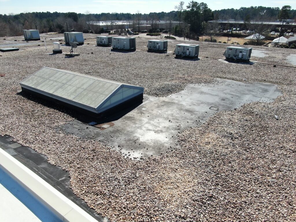 A flat commercial building roof covered with gravel, featuring several large HVAC units and a long skylight. A patch of black roofing material near the skylight may suggest recent maintenance or roof leak repair cost concerns. 