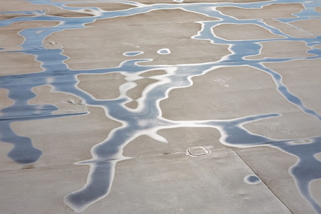 Puddles of water form irregular, reflective patterns on a smooth, light-colored concrete surface of roof