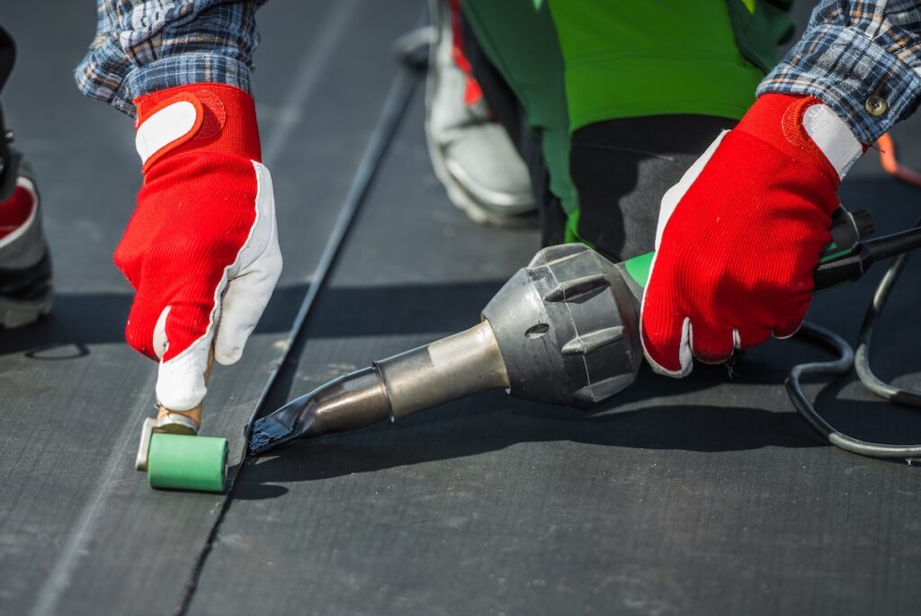 A person wearing red and white gloves uses a hot air welding tool and a small roller to seal seams on a flat black membrane during EPDM roof repair.