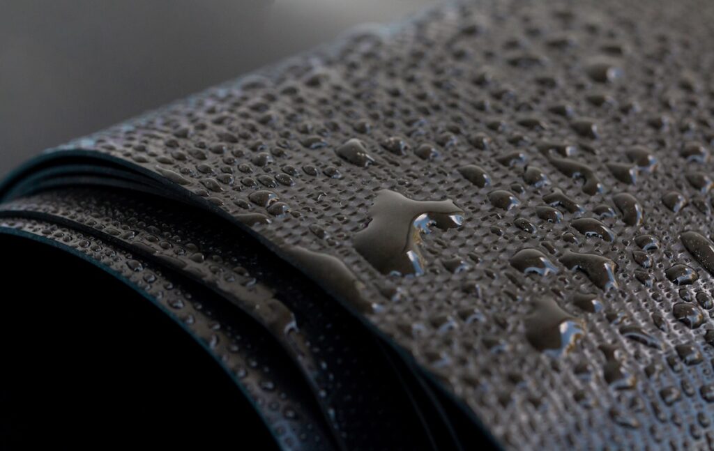 Close-up of a rolled sheet of dark, textured EPDM material with water droplets scattered across its surface, highlighting its water-resistant properties