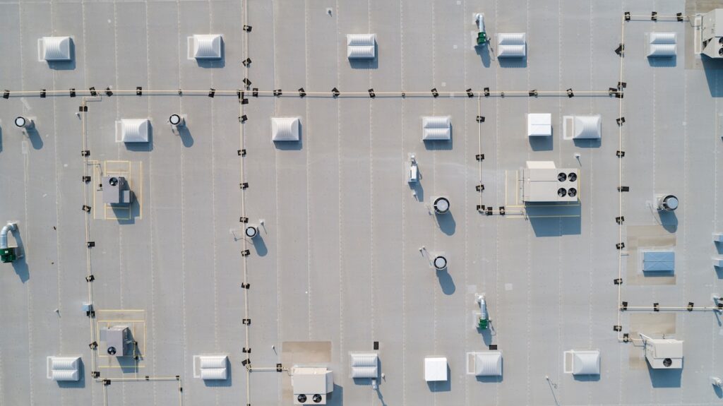 Aerial view of a large flat industrial rooftop with TPO roofing, multiple vents, HVAC units, and small rectangular structures arranged in a grid-like pattern.