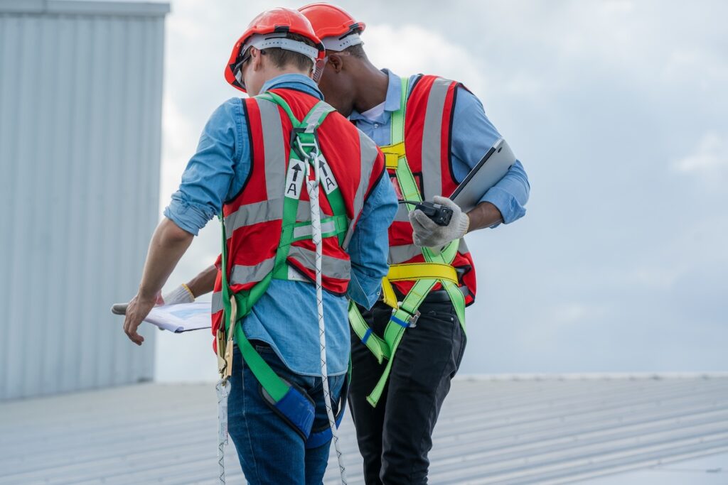 Two construction workers in safety harnesses and red vests stand on a flat roof, reviewing documents and a tablet