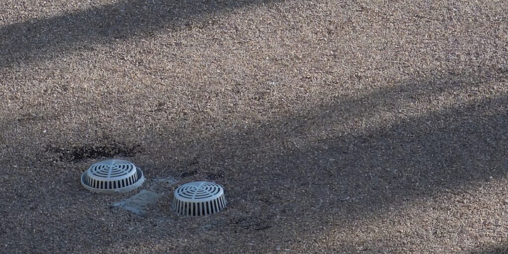 Two metal drains with slotted covers, essential parts of a commercial roof, are embedded in a gravel or pebble-covered surface