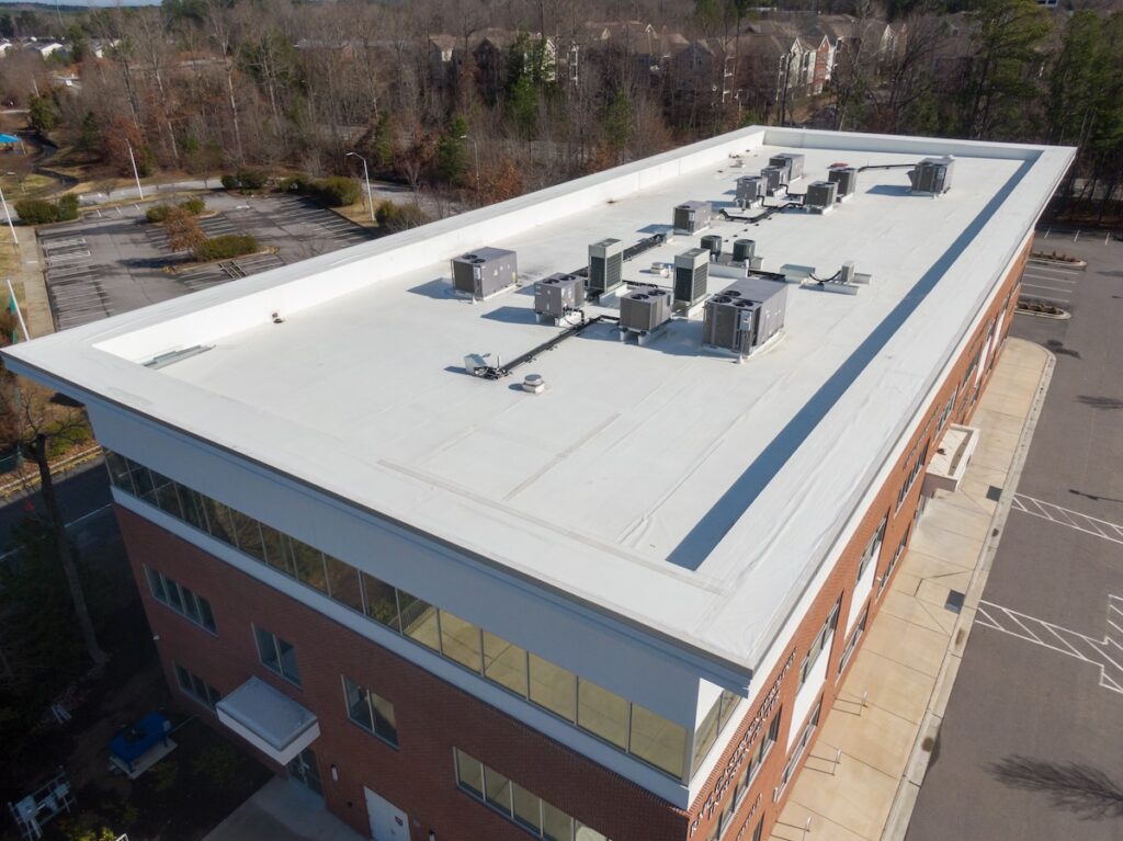 Aerial view of a flat, white-roofed commercial building with multiple HVAC units on top surrounded by parking lots, trees, and neighboring buildings.
