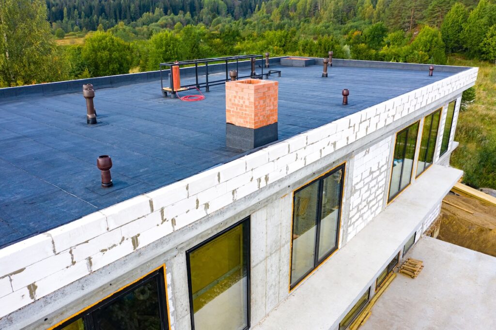 A flat-roofed building under construction with white brick walls, large windows, black roofing material, and several roof vents stands against a backdrop of green trees and forest