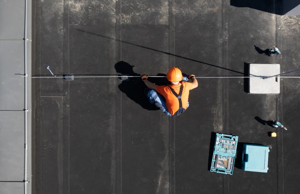 A worker wearing an orange helmet and vest is crouched on a black flat modified bitumen roof