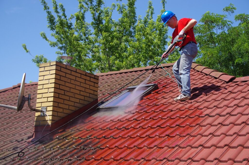 A person wearing a blue helmet and red shirt uses a power washer to clean moss off of a red tiled roof near a brick chimney and window
