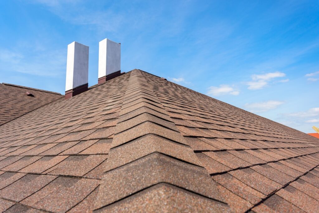 Close-up view of a roof, featuring brown asphalt shingles and two white chimneys under a blue sky with scattered clouds