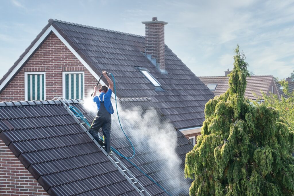 A worker in overalls stands on a ladder, pressure washing the roof of a brick house