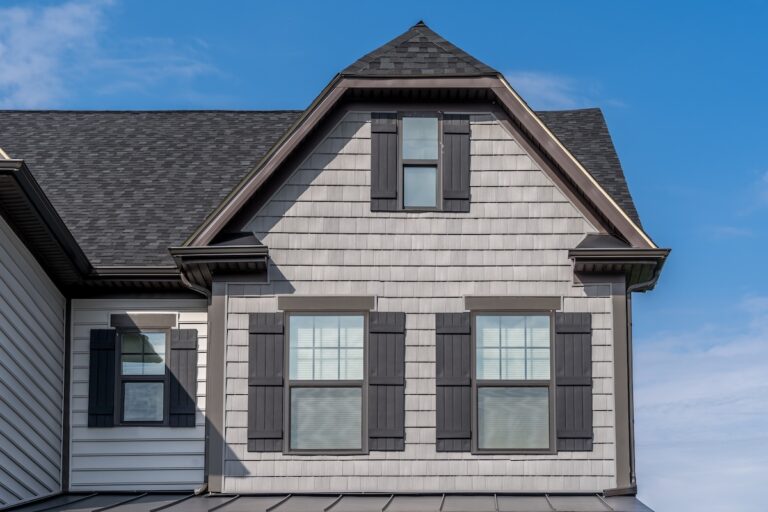 A close-up of a house exterior with light gray siding, black shutters, and three windows beneath a sharply peaked gable roof