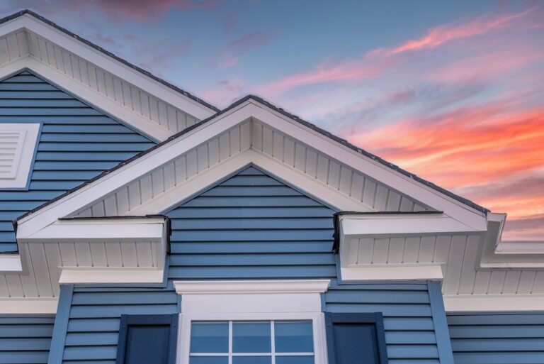 Close-up of the upper section of a blue house with white trim, featuring gabled roofs, a window with shutters, and visible fascia boards beneath the roof edges