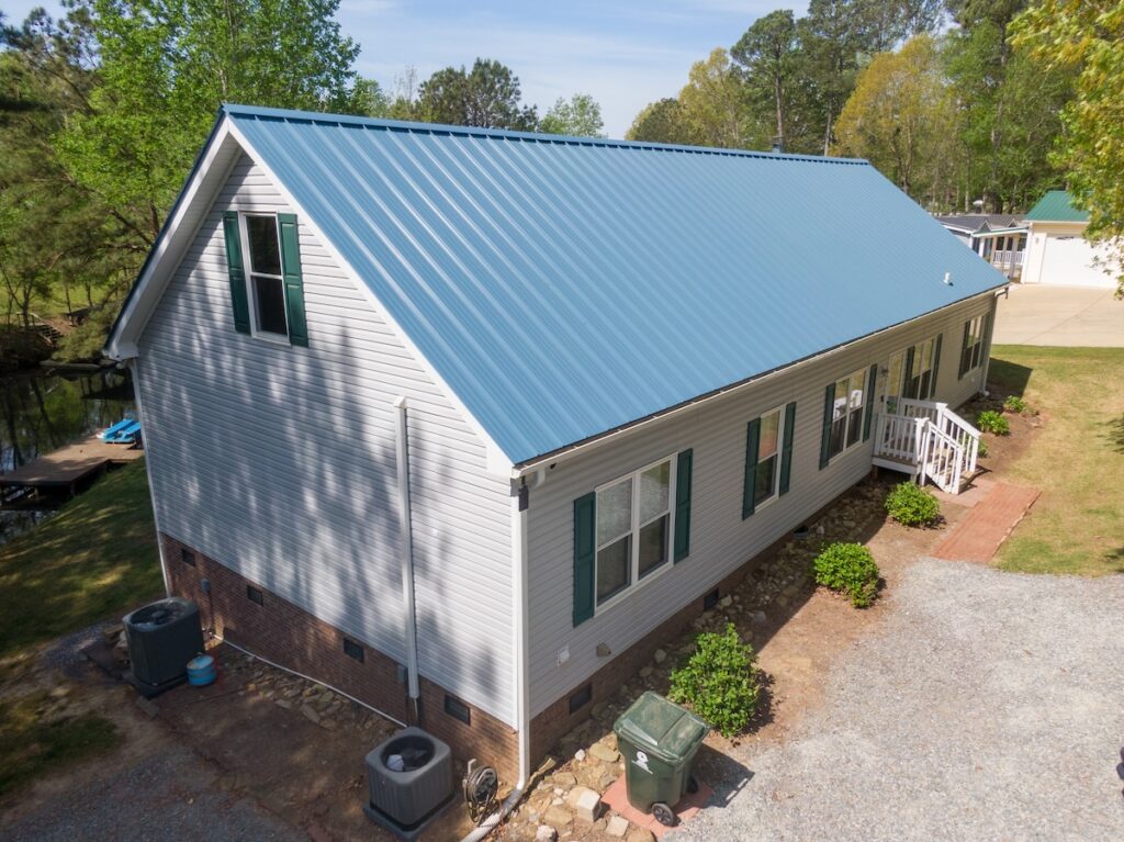 Aerial view of a light gray house with a blue metal roof, green shutters, and white trim, surrounded by trees.