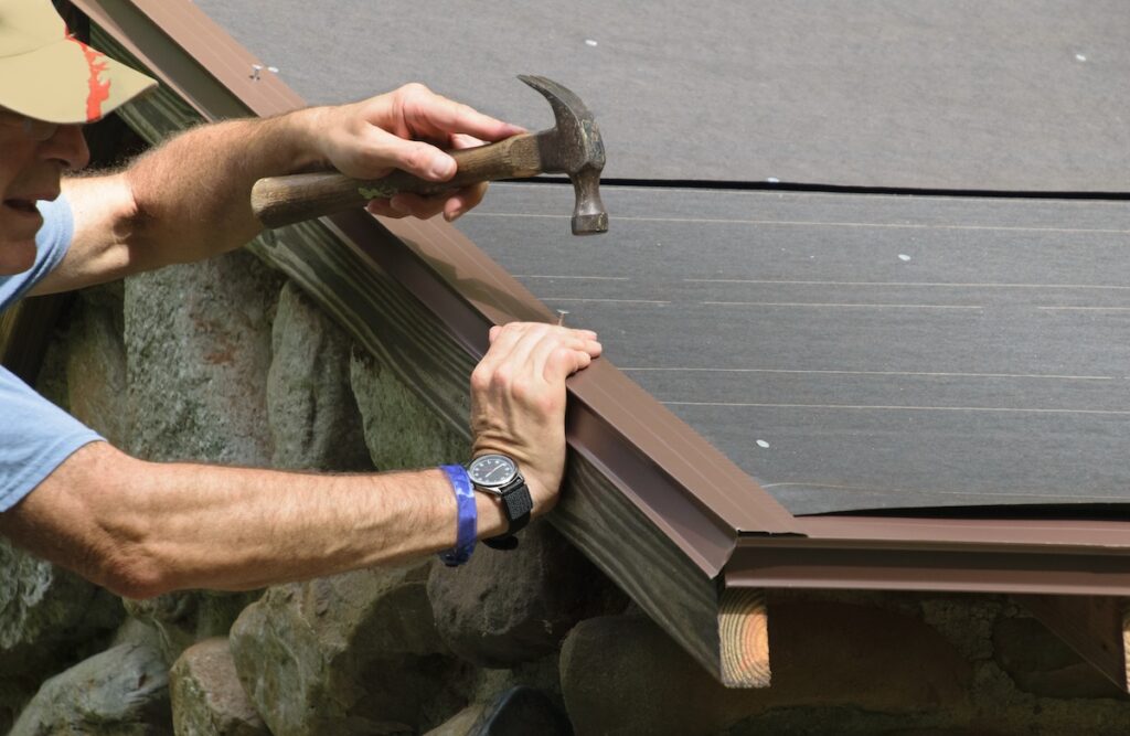 A person wearing a watch uses a hammer to nail a brown metal trim onto the edge of a roof