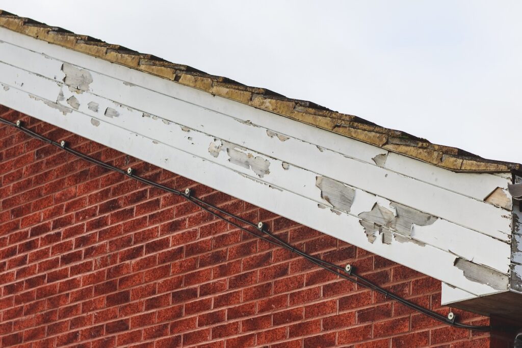 The image shows a close-up of a house's exterior, featuring a red brick wall and a white wooden fascia board with peeling paint beneath a shingle roof