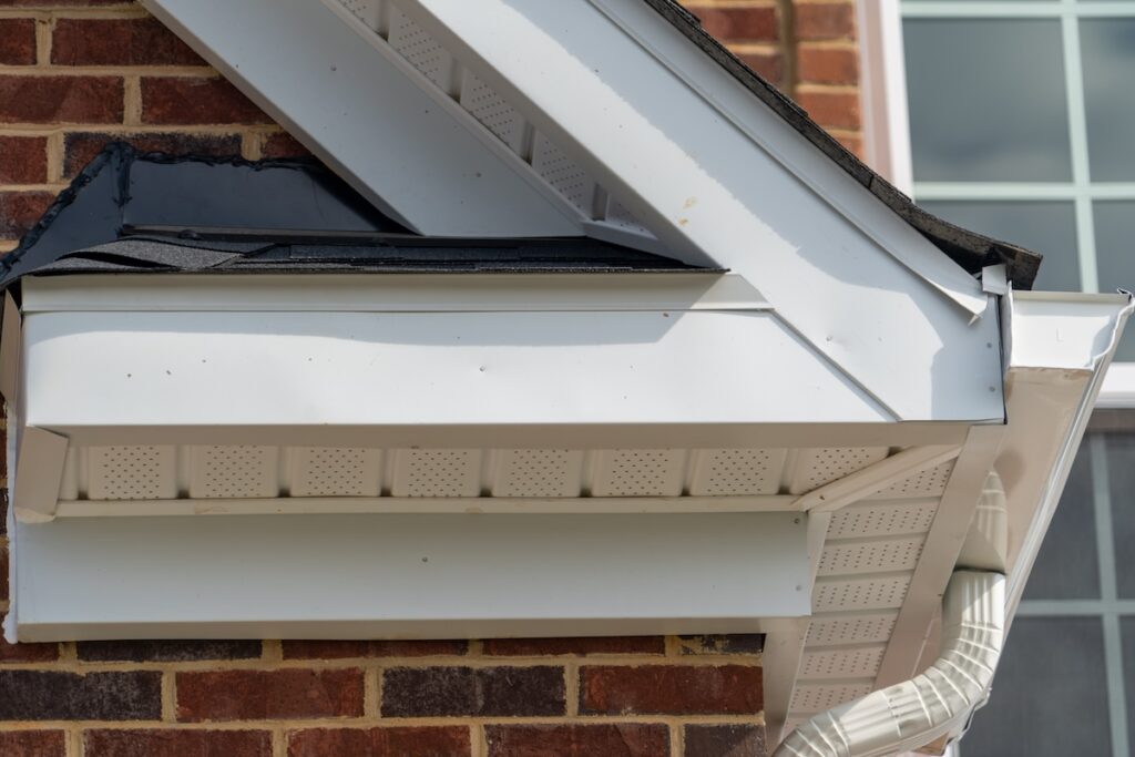 Close-up of a house corner showing a white gutter system and vented soffit on a brick wall, with an angled roof.