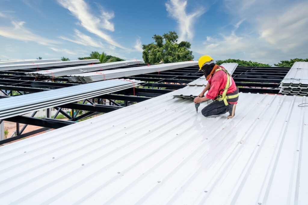 A construction worker wearing a yellow helmet and protective clothing installs white flat metal roof sheets on a large structure