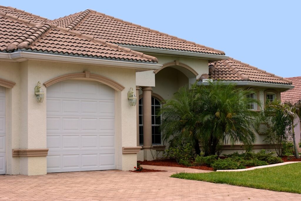A modern, single-story house with a tiled hip roof arched entryway, white garage door, palm trees, and a landscaped front yard under a clear blue sky
