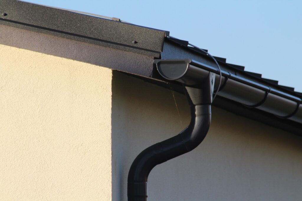 Close-up of a house corner showing a black rain gutter and downspout system attached to a beige wall under a dark roof