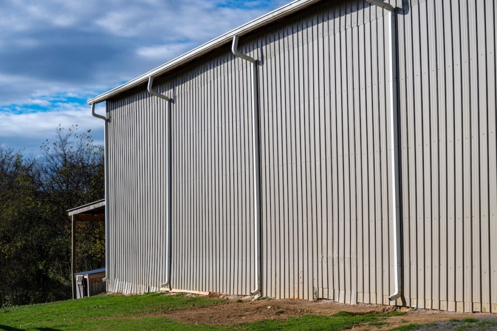 Side view of a large beige metal building with vertical corrugated panels and three white downspouts