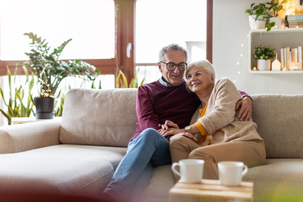 An older couple sits on a beige sofa, smiling and embracing each other in a cozy, well-lit living room with green plants and books on the shelves. Two white mugs rest on a wooden table in the foreground.