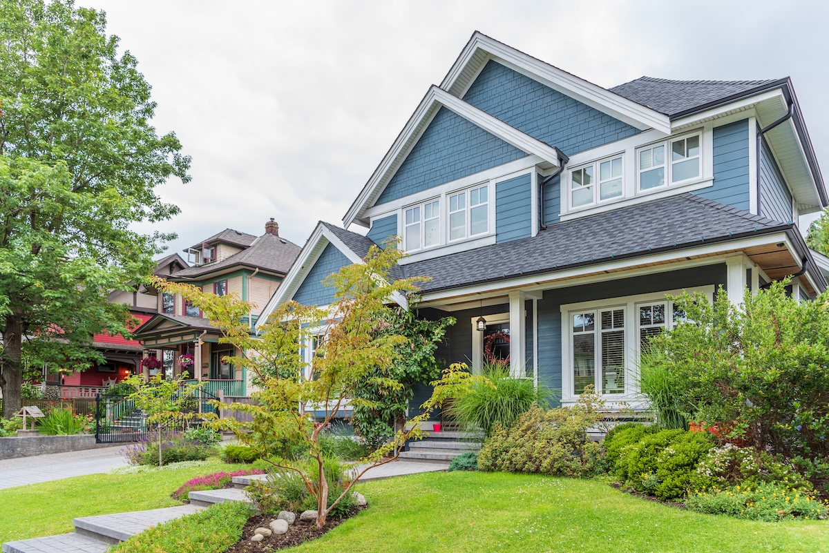 A two-story blue house with white trim, large windows, and a front porch surrounded by a well-maintained garden and green shrubs on a residential street.