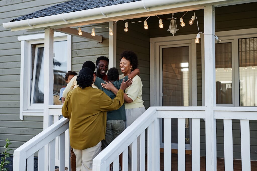 A group of people happily embrace and greet each other on the porch of a house, decorated with string lights, on a sunny day.