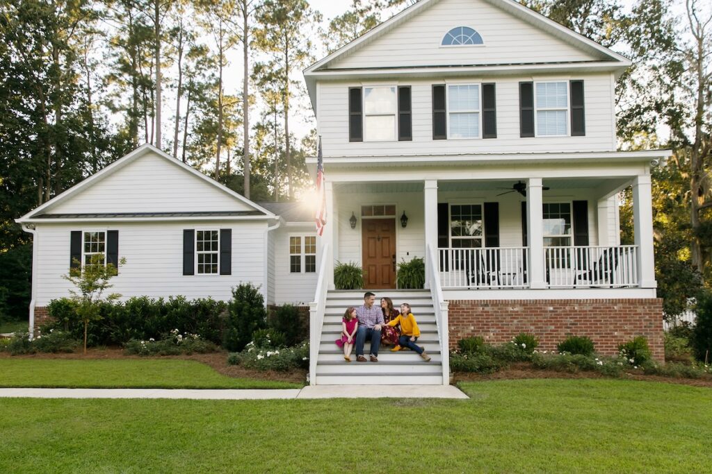 A family of four sits together on the front steps of a white two-story house with a porch, green lawn, and American flag, surrounded by trees and shrubs on a sunny day.