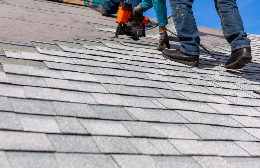 A person wearing gloves and boots uses a nail gun to install gray asphalt shingles on a sloped roof under a clear blue sky.