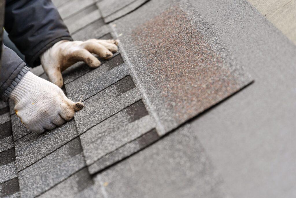 A person wearing white gloves installs or repairs gray asphalt shingles on a roof, carefully aligning the shingles for proper placement.