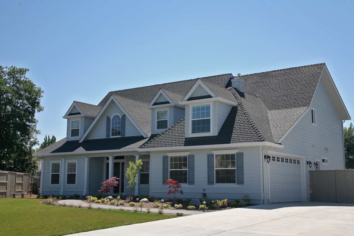 A large, modern, two-story suburban house with gray siding, white trim, multiple windows, dormers, and a neatly landscaped front yard under a clear blue sky.
