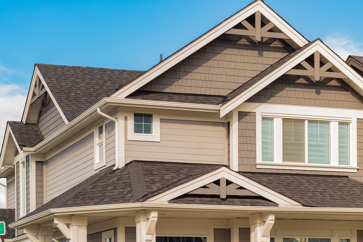 A close-up view of the upper portion of a modern two-story house with beige siding, white trim, large windows, and multiple gabled roofs under a clear blue sky.