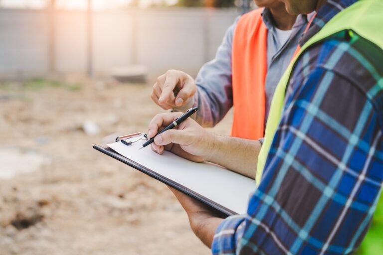Two people at a construction site, one in a plaid shirt and safety vest writing on a clipboard while another in a safety vest points at the paper.