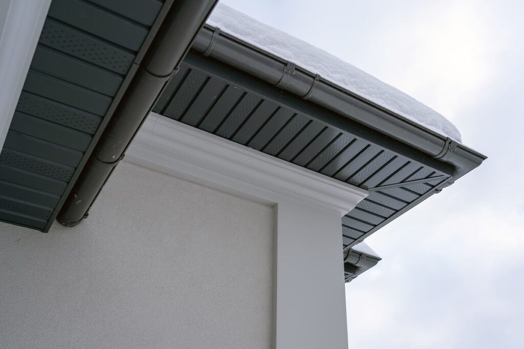 Close-up view of a building’s roof eaves with dark-colored soffit panels, rain gutter, and snow resting on the roof.