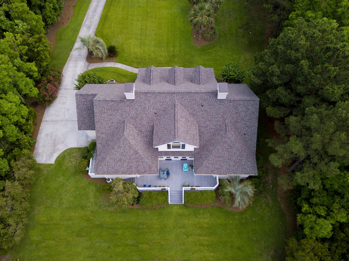 Aerial view of a large house with a shingled roof by roofing Lafayette Hill PA, surrounded by green grass, trees, and a concrete driveway leading to the rear. A front porch with outdoor furniture is visible at the entrance.