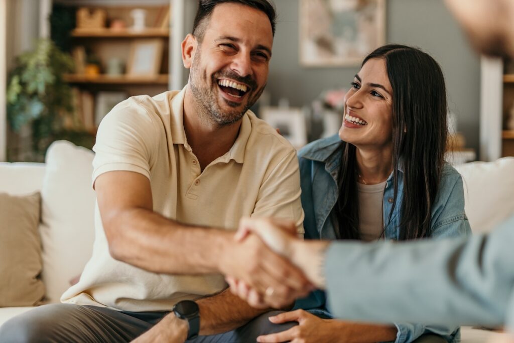 A smiling man and woman sit on a couch in a cozy living room. The man is shaking hands with someone out of frame, perhaps discussing roofing in East Norriton Township, PA, while the woman looks at him and smiles.