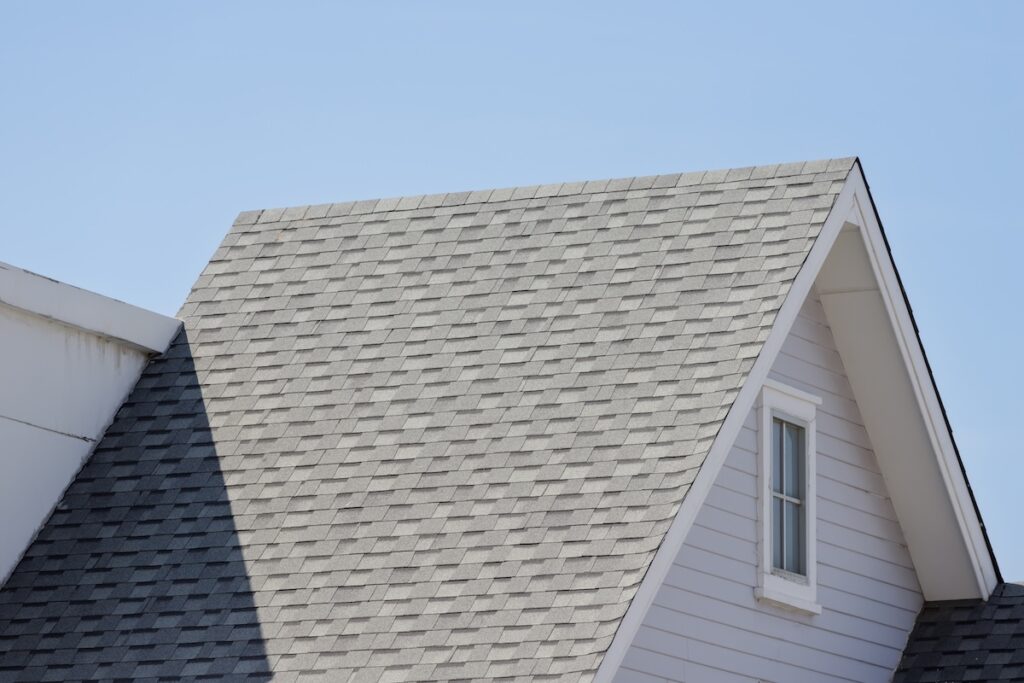 A close-up view of a house roof with light gray asphalt shingles, a sharp gable peak, and a single small window against a clear blue sky.