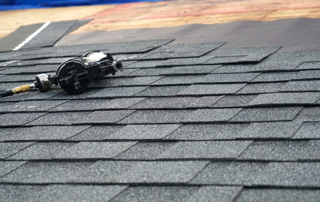 A close-up of gray asphalt shingles being installed on a roof in East Norriton Township, PA, with a pneumatic nail gun resting on top of the shingles. Perfect for showcasing quality roofing in the area.