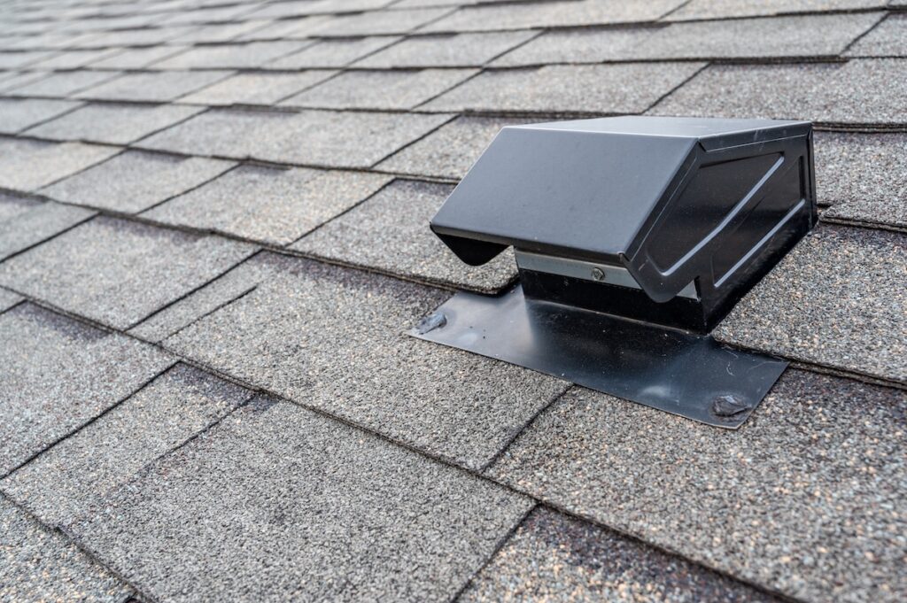 A close-up view of a black metal roof vent installed on a grey asphalt shingle roof. 