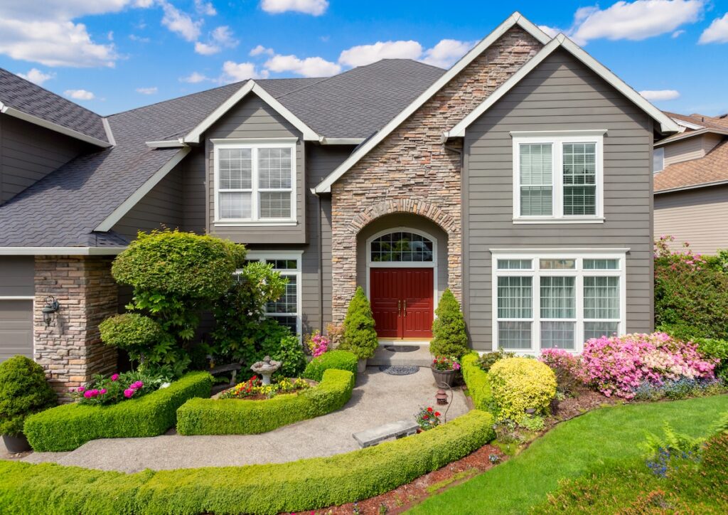 A large, modern house with gray siding, stone accents, and red double doors. The front yard features manicured shrubs, colorful flowers, and a curving walkway leading to the entrance under a bright, partly cloudy sky.
