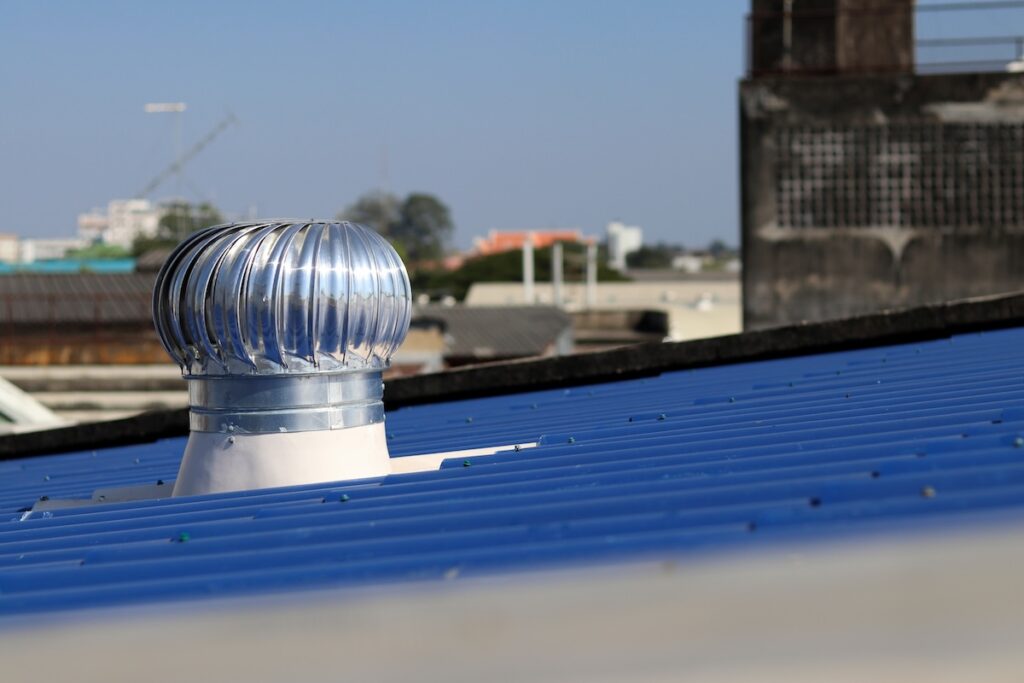 A shiny metal turbine vent is installed on a sloped blue metal roof, with blurred buildings and construction cranes visible in the background under a clear sky.