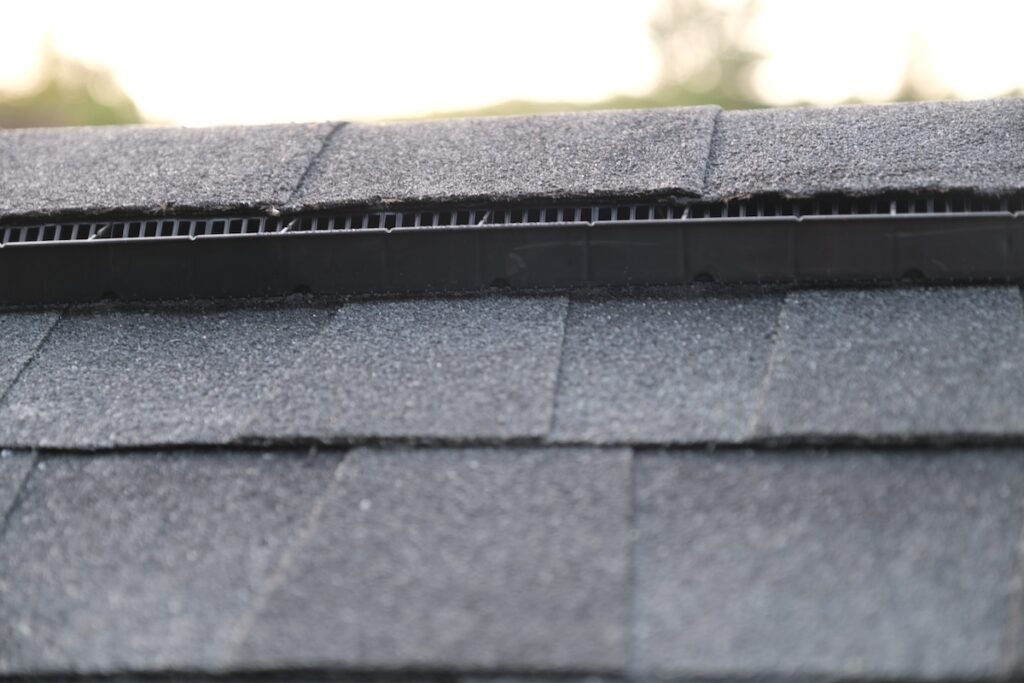 Close-up of a roof ridge vent with asphalt shingles, showing the black vent running along the peak of the roof and overlapping shingles on both sides under soft outdoor lighting.