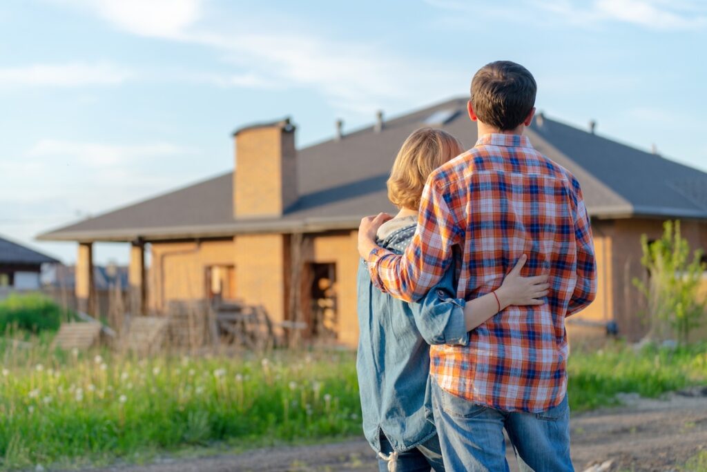 A couple stands outdoors with arms around each other, facing a newly constructed brick house on a sunny day, possibly admiring their future home