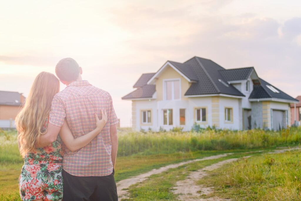 A couple stands outdoors with their arms around each other, looking at a modern yellow and white house with new roofing