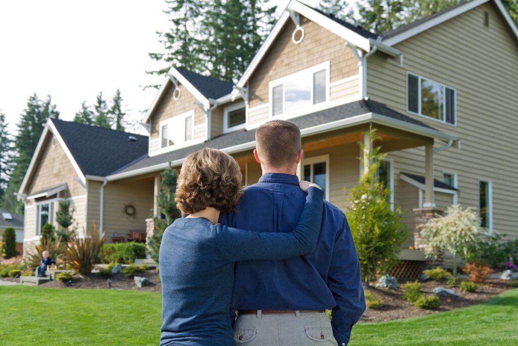 A couple stands outside with their arms around each other, looking at a modern two-story house with a well-kept yard and trees