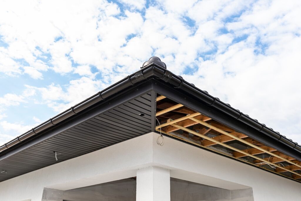 The image shows the corner of a house under construction, with one side of the roof finished and the other side exposing wooden beams and rafters.