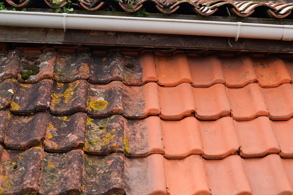 Close-up of a tiled roof showing a clear contrast between old, weathered tiles covered in moss on the left and clean, new orange tiles on the right, with a white gutter above.